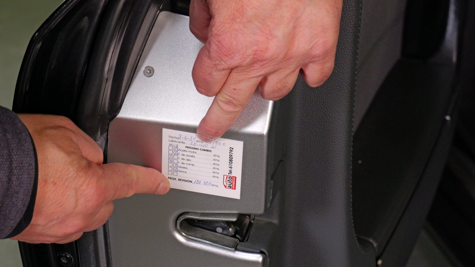 Mano colocando una pegatina de mantenimiento Autosintetico en el marco de una puerta de coche, con campos para cambios de aceite, filtros y revisiones. Pegatinas autoestáticas para talleres mecánicos, ideales para una gestión eficiente del mantenimiento